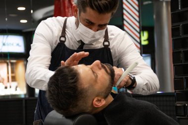 Papyonlu ve beyaz gömlekli berber müşteriyi tıraş ediyor.