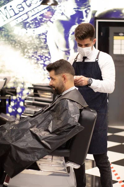 young and elegant barber with mask cuts the hair of a client with Caucasian features