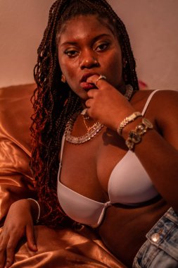 Genç Afrikalı kadın odasında yatıyor. Sahnede kahverengi ve turuncu renkler mevcuttur. Elleri dudaklarında.