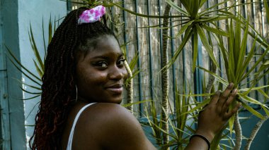 Beyaz elbiseli genç Afrikalı kadın kameraya bakarken gülümsüyor ve elini tropikal bir bitkinin üzerinde dinlendiriyor..