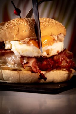 Siyah elli çocuk, yanında yağda yumurta ve siyah tepside patates kızartmasıyla lezzetli bir hamburger keser. Arka plan renkli çiçeklerden oluşuyor. Hamburger İspanya 'da yapıldı..