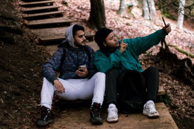 Mavi ceketli ve kapüşonlu genç yürüyüşçü, dağ kıyısındaki tahta yolda yürüyüş yapan arkadaşıyla poz veriyor.