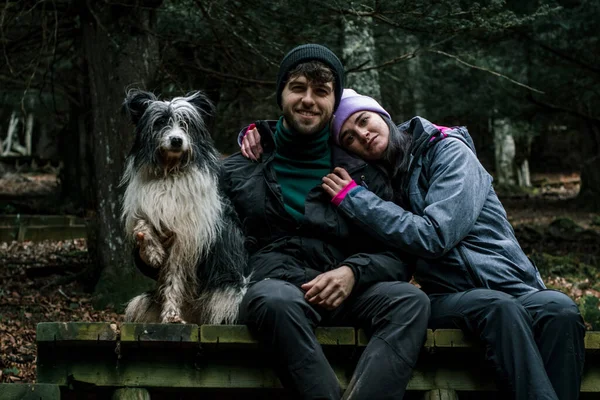 The young couple are sitting on a wooden plank road in the forest. Next to them is their shepherd dog.