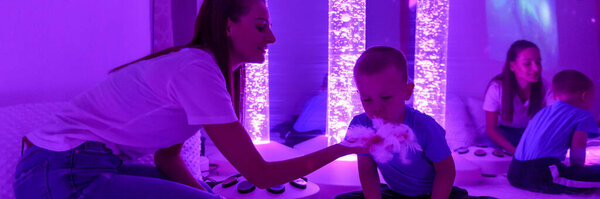Child with intellectual disability and his therapist in sensory stimulating room, snoezelen. Autistic child interacting with therapist during therapy session. Web banner.