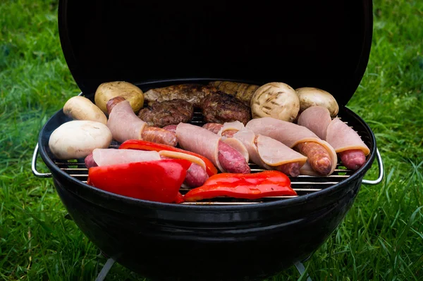 Vista de alto ângulo de bifes suculentos, hambúrgueres, salsichas e legumes cozinhando em um churrasco sobre as brasas em um gramado verde ao ar livre — Fotografia de Stock