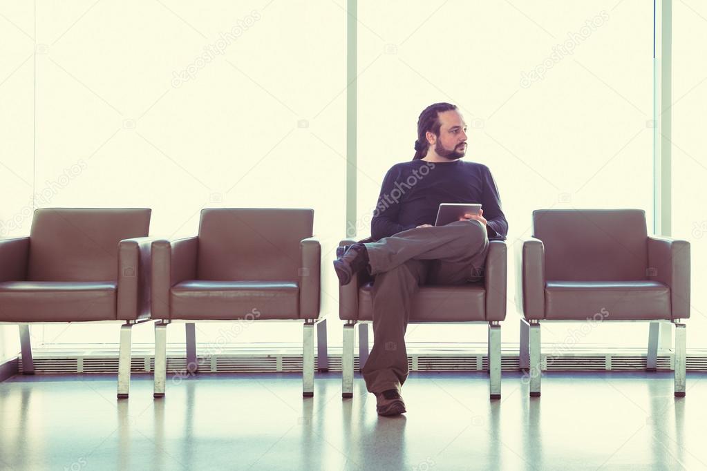 Handsome young man with dreadlocks using his digital tablet pc at an airport lounge, modern waiting room, with backlight.