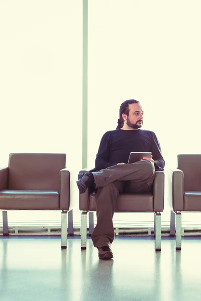 Handsome young man with dreadlocks using his digital tablet pc at an airport lounge, modern waiting room, with backlight.