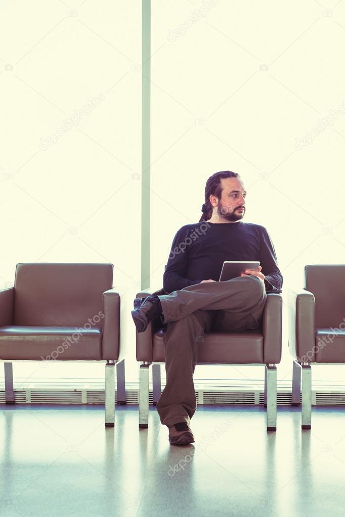 Handsome young man with dreadlocks using his digital tablet pc at an airport lounge, modern waiting room, with backlight.