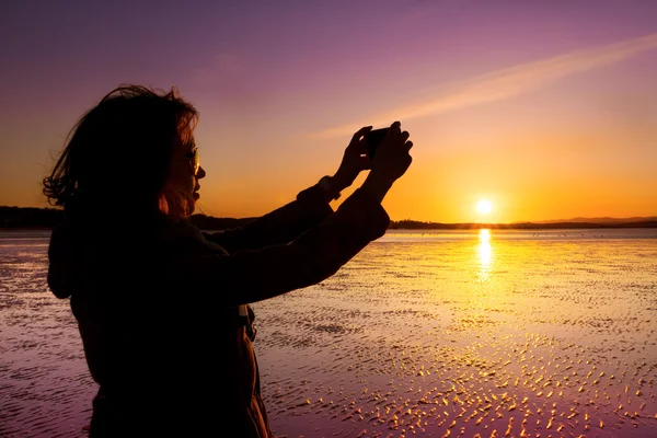 Mulher bonita tirando foto de si mesma, selfie, em uma praia durante o pôr do sol . — Fotografia de Stock