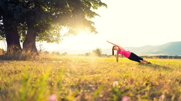 Nuori urheilullinen nainen harjoittelee joogaa niityllä auringonlaskun aikaan, kuva linssin soihdulla — kuvapankkivalokuva