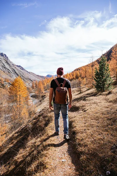 Young boy on his back wearing a black t-shirt and a red wool cap and carrying a brown leather backpack walking in the mountains