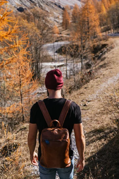 Young boy on his back wearing a black t-shirt and a red wool cap and carrying a brown leather backpack walking in the mountains