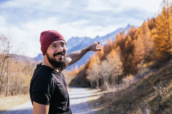 Young guy with glasses and a beard dressed in a black T-shirt and a red hat pointing to the mountains