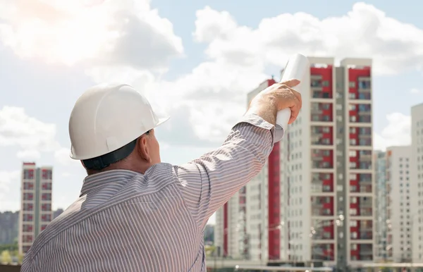 Un ingeniero con casco blanco apuntando con planos en la mano sobre edificios — Foto de Stock