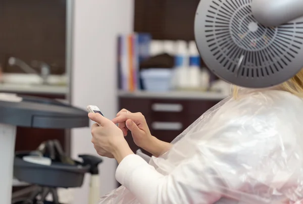 Eine blonde Frau surft mit ihrem Smartphone im Haarstudio im Internet — Stockfoto