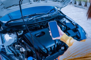 Motor dairesine tepeden bakan kaputlu bir arabanın önünde duran bir kadın akıllı telefon uygulaması aracılığıyla oto tamir merkezine ulaşmak için telefonu kullanıyor.