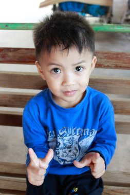 Yakışıklı Asyalı çocuk kameraya bak. Mavi gömlekli, siyah pantolonlu, salıncakta oturuyor. Elini kaldırıp parmağını kaldırdığında arka plan bulanıklaşıyor..