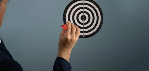 In the right hand of a man in a suit, there is a red dart that is being pinned into the center of a circular target on the wall, The goal of concep is to achieve it.
