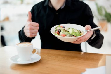 Bu salata gerçekten güzel. Adam baş parmağıyla balık yeşili ve avokado salatası tutuyor. Masada bir fincan nefis bembeyaz kahve. Manzarayı kes. Sağlıklı gıda konsepti. Siyah gömlek. Akıllı biçim.