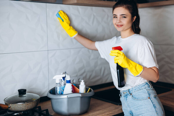 Smiling housekeeper in protective yellow gloves cleaning kitchen with detergents.