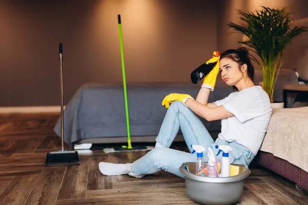 Mujer cansada en camiseta blanca con limpieza de pelo oscuro en guantes de goma amarilla para la protección de las manos y cubo con artículos de limpieza en el hogar — Foto de Stock