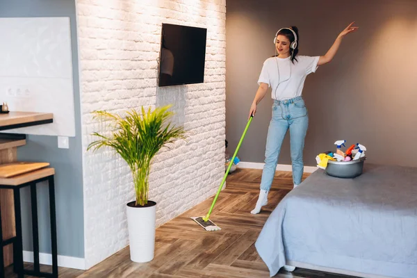 Foto de larga duración de la mujer alegre feliz está lavando el suelo en la habitación con fregona divertirse. Imagínese de ama de casa bailando en auriculares mientras limpia en interiores. — Foto de Stock