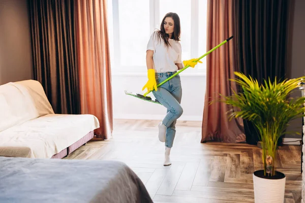 Beautiful young woman in yellow gloves dancing with mop while cleaning the living room. — Stock Photo, Image