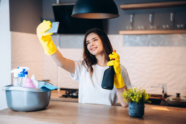 Young happy cute woman in yellow gloves is taking selfie while cleaning her kitchen at home with detergents.