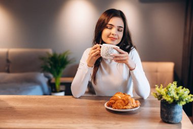Siyah, parlak saçlı, kahvaltı sırasında kahve içen genç bir kadın. Güzel esmer kızın ev portresi sabahları kruvasan yiyip çayın tadını çıkarıyor.