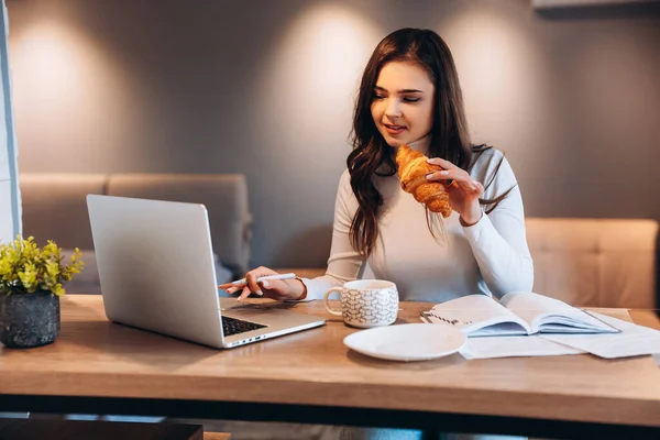 Внештатная студентка использует ноутбук, сидя дома. Молодая женщина, сидящая на кухне и работающая онлайн на ноутбуке. Красивая женщина пьет чай кофе во время работы — стоковое фото