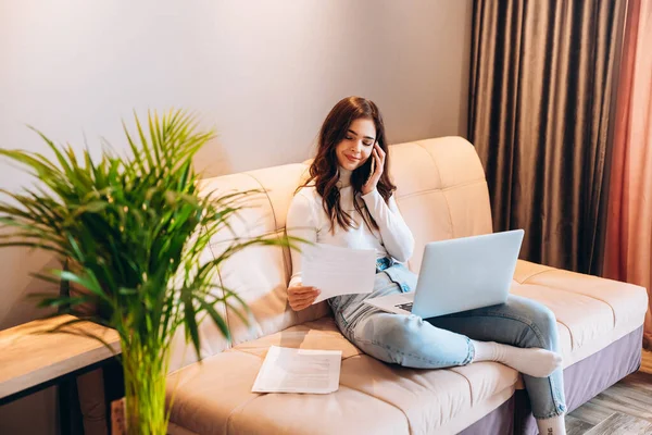 Молодая студентка-фрилансер, сидя дома, пользуется ноутбуком. Молодая женщина сидит на диване и работает онлайн на ноутбуке. Красивая женщина разговаривает по телефону во время учебы — стоковое фото