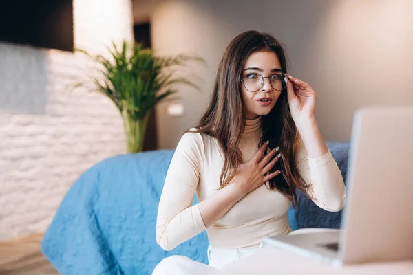 Menina encantadora em óculos está trabalhando em laptop indoor e, provavelmente, ela cometeu um erro ou tem más notícias. Freelancer feminino trabalhando em casa. Estudante interior. — Fotografia de Stock