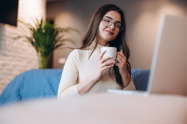 Любимая девушка в очках наслаждается чашкой чая и кофе, работая на ноутбуке в помещении. Женщина-фрилансер, работающая из дома — стоковое фото