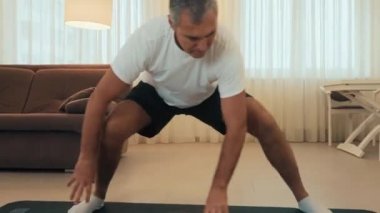 Yaşlı adam yan atlama egzersizi yapıyor. Formda vücut ısısı olan son sınıf erkek atlet, evde bacak esnetme egzersizi yapıyor. Kendinize iyi bakın. Arka planda modern ışık odası. Yaşlı spor konsepti