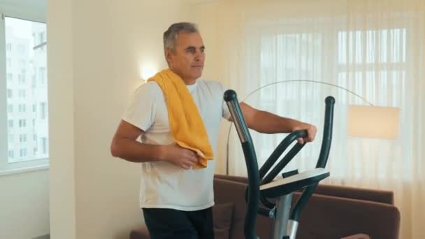 Mature happy man exercising on elliptical trainer and dries the sweat with a towel Workout at home. Cardio training using cross-trainer. White living room on background. — Stock Video