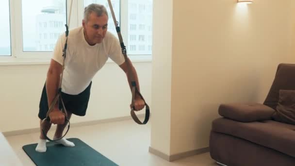 Зрелый человек делает перекресток отжимания с фитнес-ремнями для тренировки подвески. Счастливый пожилой мужчина делает упражнения с трх. Полная длина. Тренировка дома. Активный и здоровый образ жизни — стоковое видео