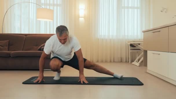 Starší muž dělá postranní výpad. Senior samec sportovec s kondicí tělo zahřívání, dělá nohou protáhnout cvičení doma. Dávejte na sebe pozor Moderní světelné místnosti na pozadí. — Stock video