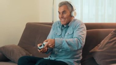 Yetişkin adam konsolda oynuyor. Hafta sonu konseptinde iyi eğlenceler. Kulaklıklı konsantre bir adam kahverengi koltukta oturuyor, oyun kumandasını tutuyor ve video oyunları oynuyor..