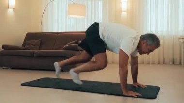 Yaşlı bir adam siyah yoga minderi üzerinde dağcı egzersizleri yapıyor. Sabah sporu. Arka planda modern oturma odası. Plastik su şişesi. Sıkı çalışma. Evde antrenman. Yaşlı spor konsepti.