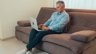 Orta yaşlı bir adam haber okumak ya da e-postalara cevap vermek için modern dizüstü bilgisayar kullanıyor. Mavi gömlek ve kot pantolon. Modern teknoloji kavramı.