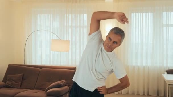 Warm up first Cheerful senior man stretching his arms, smiling and looking at camera. Morning workout on yoga mat. Strong man doing tilts of the body to the side at home. — Stock Video