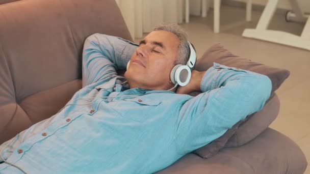 Descanse en casa después del trabajo durante la cuarentena. Hombre maduro con el pelo gris se encuentra en el sofá y escucha música utiliza auriculares blancos o auriculares. Tiempo libre. Concepto de tecnología moderna. — Vídeos de Stock