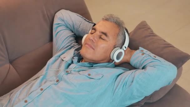 Odpoczynek w domu po pracy podczas kwarantanny. Dojrzały mężczyzna z siwymi włosami leży na kanapie i słucha muzyki używa białych słuchawek lub słuchawek. Czas wolny. Koncepcja nowoczesnej technologii. — Wideo stockowe