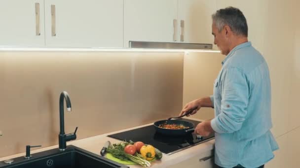 Koncentreret mand i afslappet tøj står ved ovnen og gryderetter grøntsager i en stegepande. Moden mand forbereder sund mad til middag for sin familie. Sund mad koncept – Stock-video