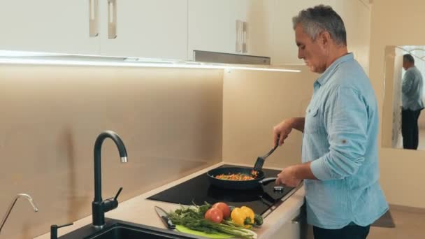 Felnőtt férfi egészséges ételt készít a családjának vacsorára. Koncentrált férfi alkalmi ruhákban áll a tűzhely mellett, és zöldségeket pörkölt egy serpenyőben. Egészséges élelmiszer-koncepció — Stock videók