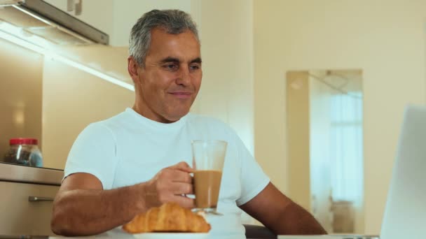 Hombre maduro disfrutando de su capuchino y delicioso croissant. Concepto de rutina matutina. Un freelancer masculino está disfrutando de su dulce desayuno, mirando al monitor del portátil y feliz de haber encontrado un cliente. — Vídeos de Stock