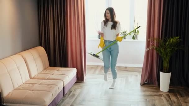 La joven limpia en casa. Morena modelo femenina está bailando y cantando con una fregona mientras limpia la casa. — Vídeo de stock