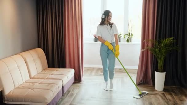 Een jonge vrouw maakt thuis schoon. Brunette vrouwelijk model is luisteren naar muziek en zingen tijdens het schoonmaken van het appartement of huis. — Stockvideo