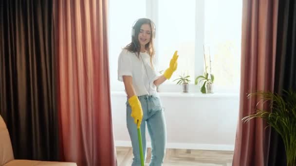 La joven limpia en casa. Morena modelo femenina está escuchando música y cantando mientras limpia el apartamento o la casa. — Vídeos de Stock
