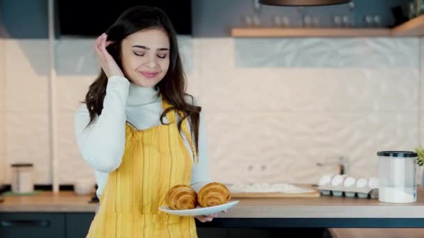 Joven mujer divertida untó su cara con harina mientras cocinaba. Ella no sabe que toda la harina, modelo femenina es una encantadora sonriendo a la cámara y mostrando croissants recién hechos. — Vídeos de Stock